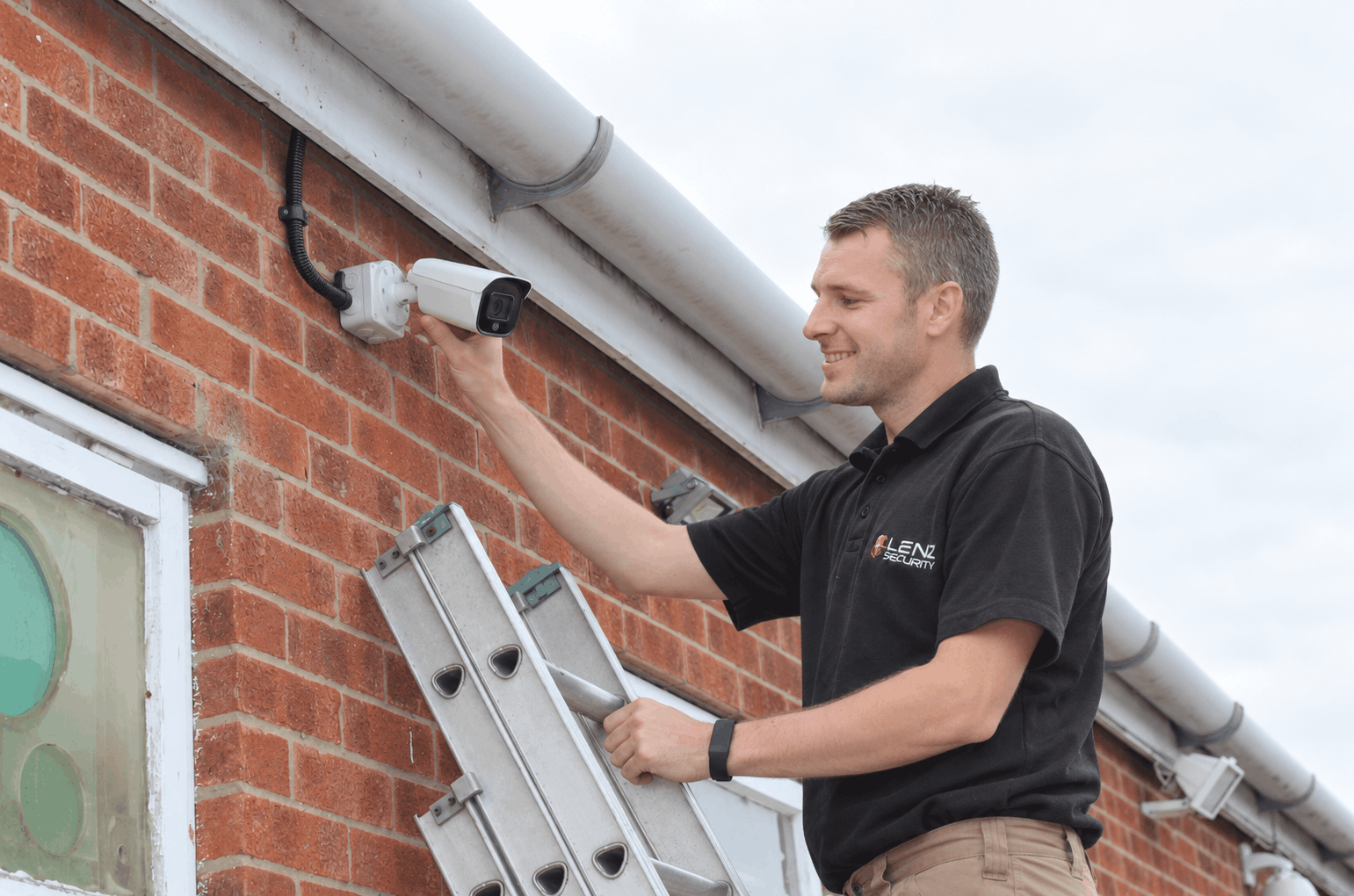 CCTV engineer inspects CCTV camera while on ladder in front of a brick house for “How often should CCTV be serviced” blog by Lenz Security